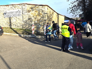 Na zdjęciu policjant rozmawia z mężczyzną, który trzyma za rękę dziewczynkę. Znajdują się obok cmentarza.