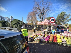 Na zdjęciu widzimy zaparkowany radiowóz przy cmentarzu oraz policjanta który stoi obok niego.