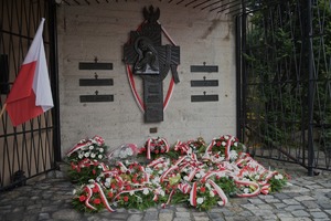 Na zdjęciu widzimy złożone kwiaty pod tablicą pamiątkową, widoczne są flagi biało-czerwone.