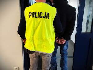 Na zdjęciu policjant w kamizelce z napisem POLICJA wyprowadza zatrzymanego, który ma założone kajdanki z policyjnego aresztu.