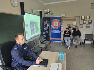 Na zdjęciu umundurowany policjant siedzi za biurkiem w klasie . W oddali dwóch mężczyzn siedzi na krześle.