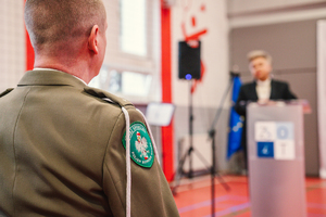 Na zdjęciu widzimy fragment munduru funkcjonariusza straży granicznej oraz kobietę przemawiającą na mównicy.