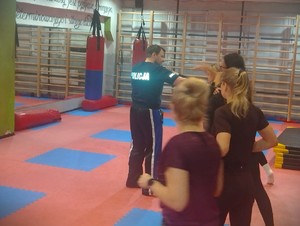 Na zdjęciu mężczyzna w koszulce policyjnej uczy kobiety na sali treningowej samoobrony.