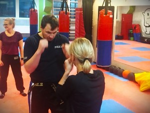 Na zdjęciu mężczyzna w koszulce policyjnej uczy kobietę jak trzymać gardę.