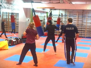 Na zdjęciu mężczyzna w koszulce policyjnej patrzy jak kobiety wykonują ćwiczenia na sali treningowej.
