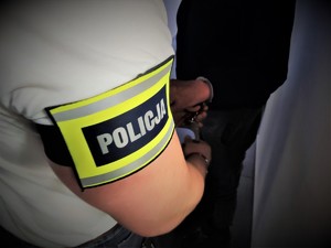 Na zdjęciu zbliżenie na ramię policjanta, który ma założoną opaskę z napisem POLICJA podczas zakładania kajdanek zatrzymanemu.