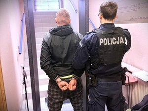 Na zdjęciu policjant doprowadza zatrzymanego mężczyznę, który ma założone kajdanki na ręce trzymane z tyłu