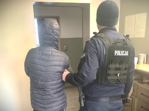 Na zdjęciu znajduje się policjant trzymający za ramię zatrzymanego, który ma założone kajdanki na ręce trzymane z przodu.