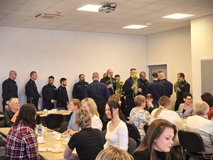 Zdjęcie grupowe. Policjanci w mundurach stoją z kwiatkami, a kobiety siedzą przy stołach.