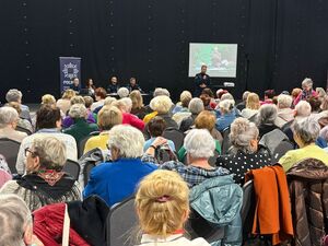 Na zdjęciu policjant występuje przed grupą seniorów na hali sportowej.
