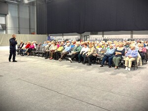 Na zdjęciu policjant występuje przed grupą seniorów na hali sportowej.