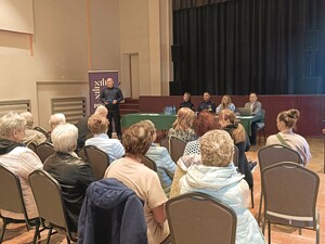 Zdjęcie grupowe podczas debaty na temat bezpieczeństwa seniorów na sali konferencyjnej. Widzimy seniorów, policjantów i zaproszonych prelegentów. Występuje przed seniorami policjant z mikrofonem.