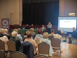 Zdjęcie grupowe podczas debaty na temat bezpieczeństwa seniorów na sali konferencyjnej. Widzimy seniorów, policjantów i zaproszonych prelegentów. Występuje zaproszona prelegentka z prokuratury.