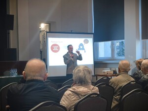 Zdjęcie grupowe podczas debaty na temat bezpieczeństwa seniorów na sali konferencyjnej. Widzimy seniorów, oraz prelegenta występującego przed seniorami.