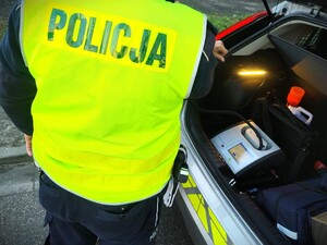 Na zdjęciu zbliżenie na policjanta w kamizelce odblaskowej i alkomat, który znajduje się w bagażniku radiowozu.