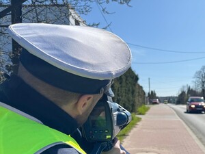 Na zdjęciu policjant przy drodze podczas mierzenia prędkości urządzeniem pomiarowym.